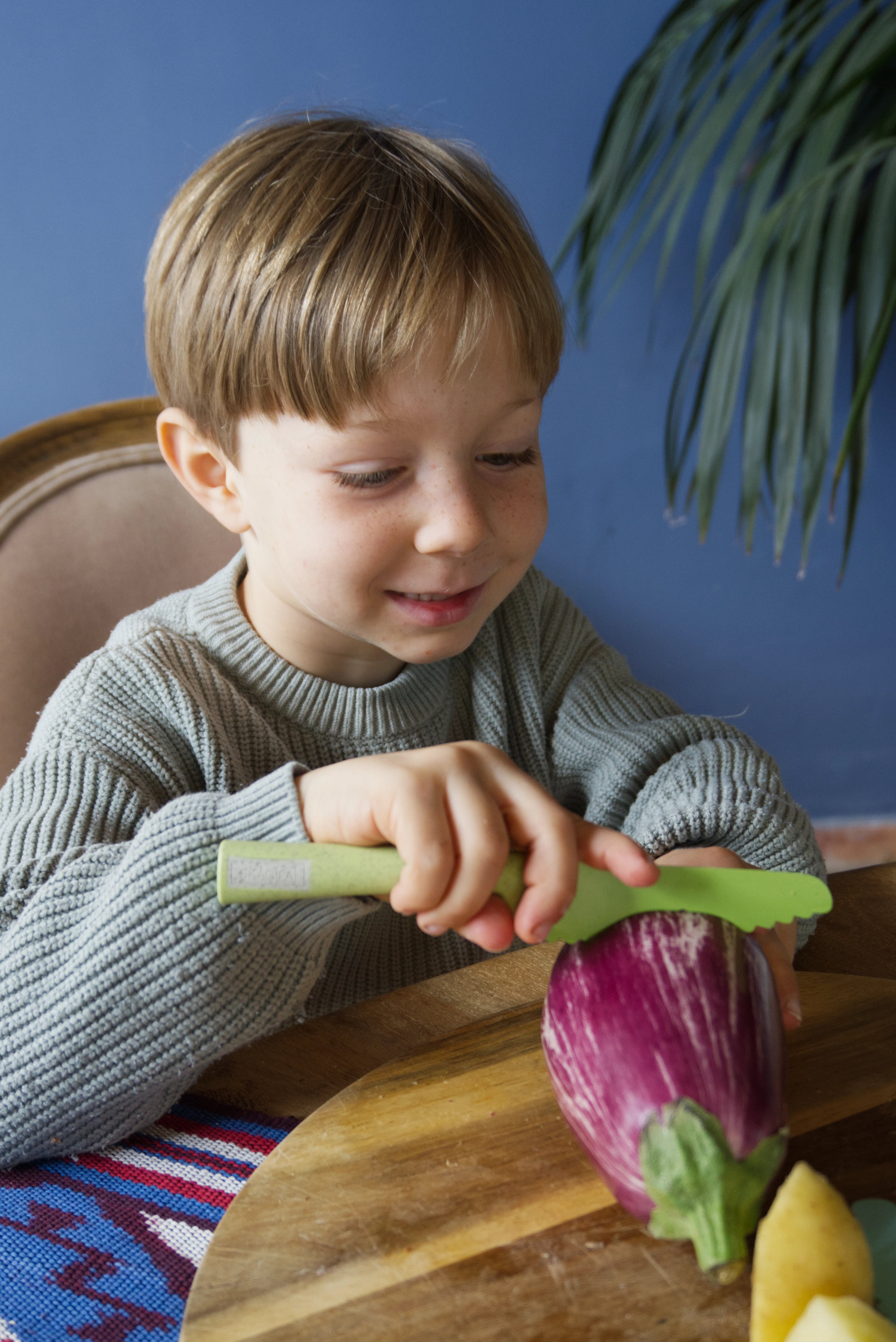 Child holding a toy eggplant on a wooden table with a blue wall in the background, Klyv classic knife for 3 years old Scandinavian Design