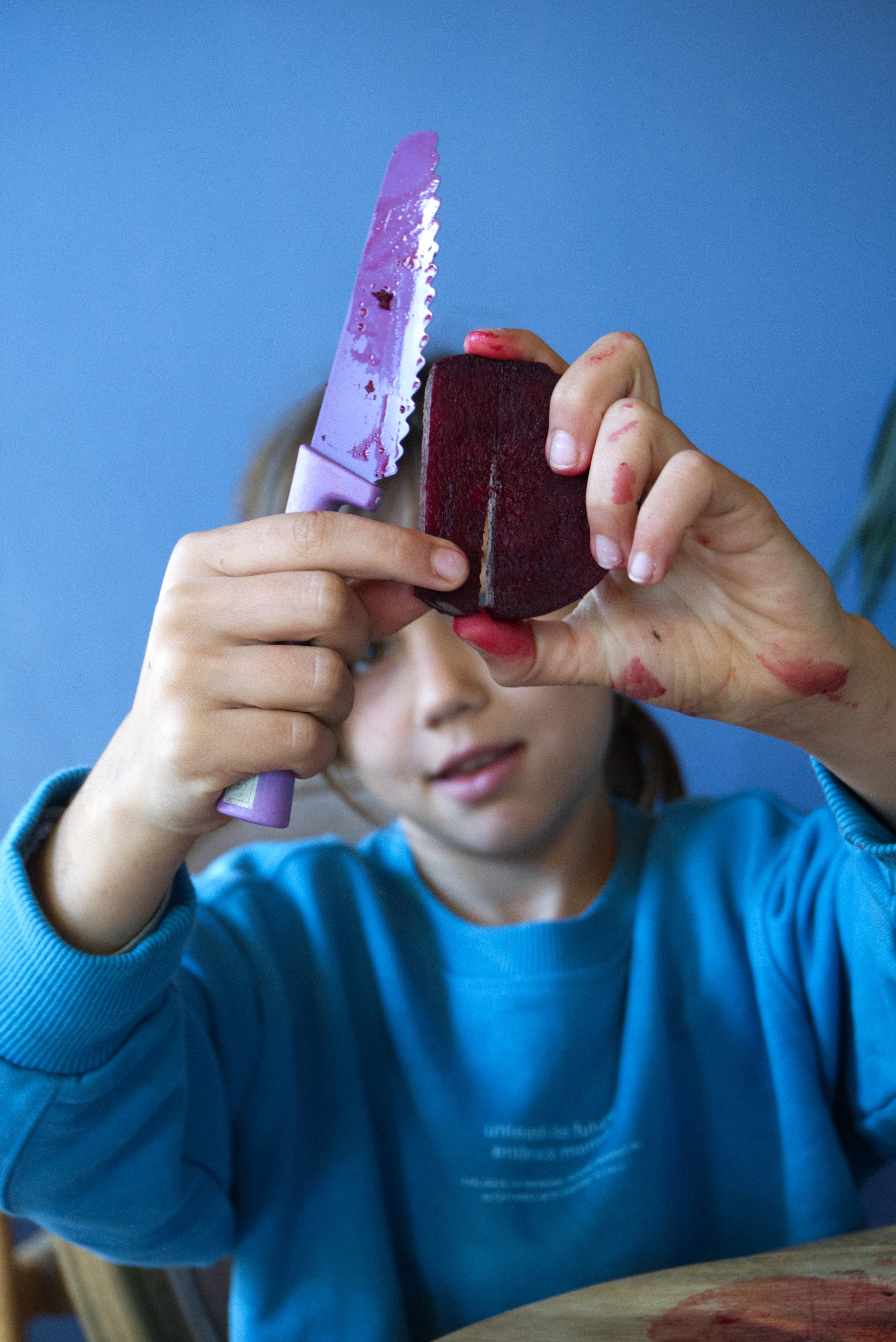 Girl holding a purple kids knife and a piece of beets against a blue background, Klyv classic knife for 3 years old Scandinavian Design