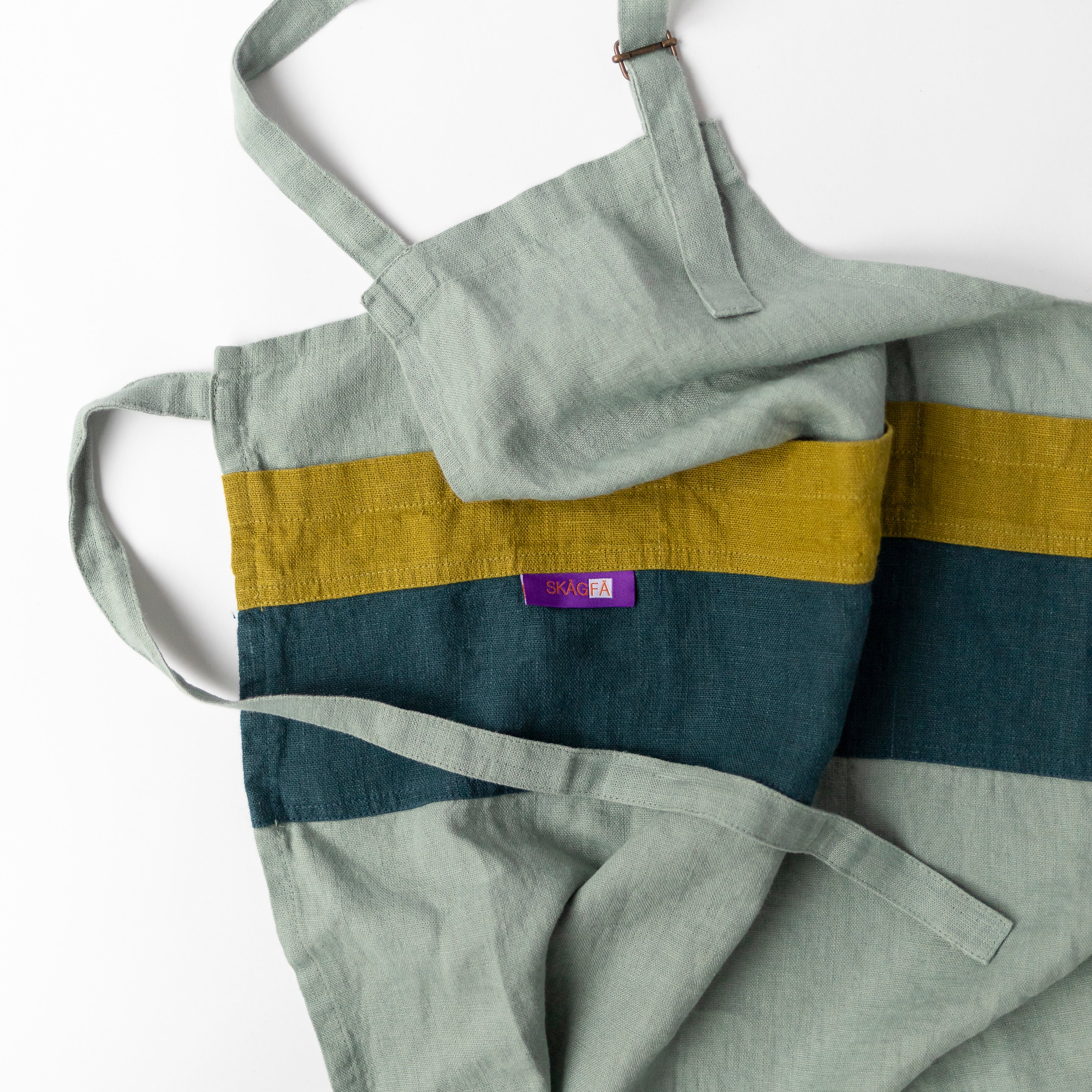 Child in a kitchen wearing 'The Förkläde' linen chef apron by SKÅGFÄ, showcasing adjustable neck strap and deep pockets, designed in Spain and handmade in Europe, perfect for culinary adventures. The apron features chic simplicity with 100% OEKO-TEX certified stone-washed linen, promoting sustainable fashion and functionality for young chefs blue navy pink peach salmon sage green forest