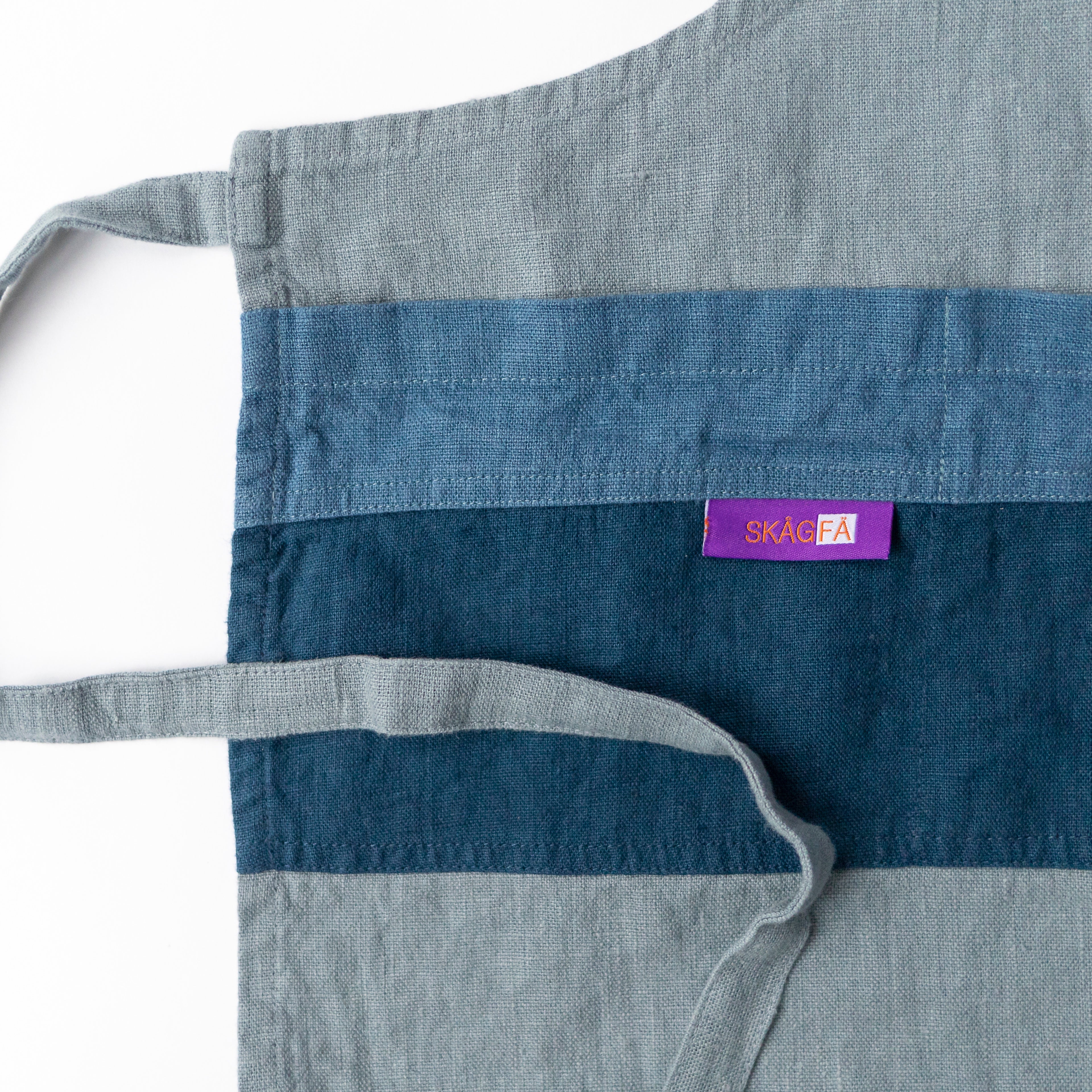 Child in a kitchen wearing 'The Förkläde' linen chef apron by SKÅGFÄ, showcasing adjustable neck strap and deep pockets, designed in Spain and handmade in Europe, perfect for culinary adventures. The apron features chic simplicity with 100% OEKO-TEX certified stone-washed linen, promoting sustainable fashion and functionality for young chefs blue navy pink peach salmon sage green forest