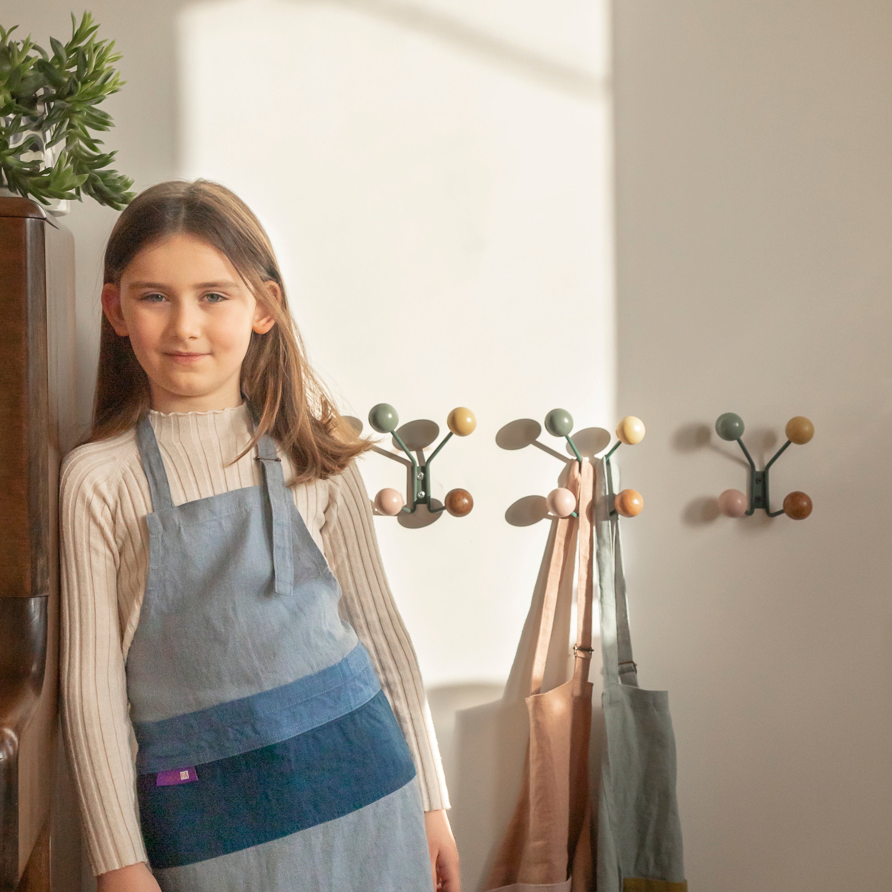 Child in a kitchen wearing 'The Förkläde' linen chef apron by SKÅGFÄ, showcasing adjustable neck strap and deep pockets, designed in Spain and handmade in Europe, perfect for culinary adventures. The apron features chic simplicity with 100% OEKO-TEX certified stone-washed linen, promoting sustainable fashion and functionality for young chefs blue navy pink peach salmon sage green forest