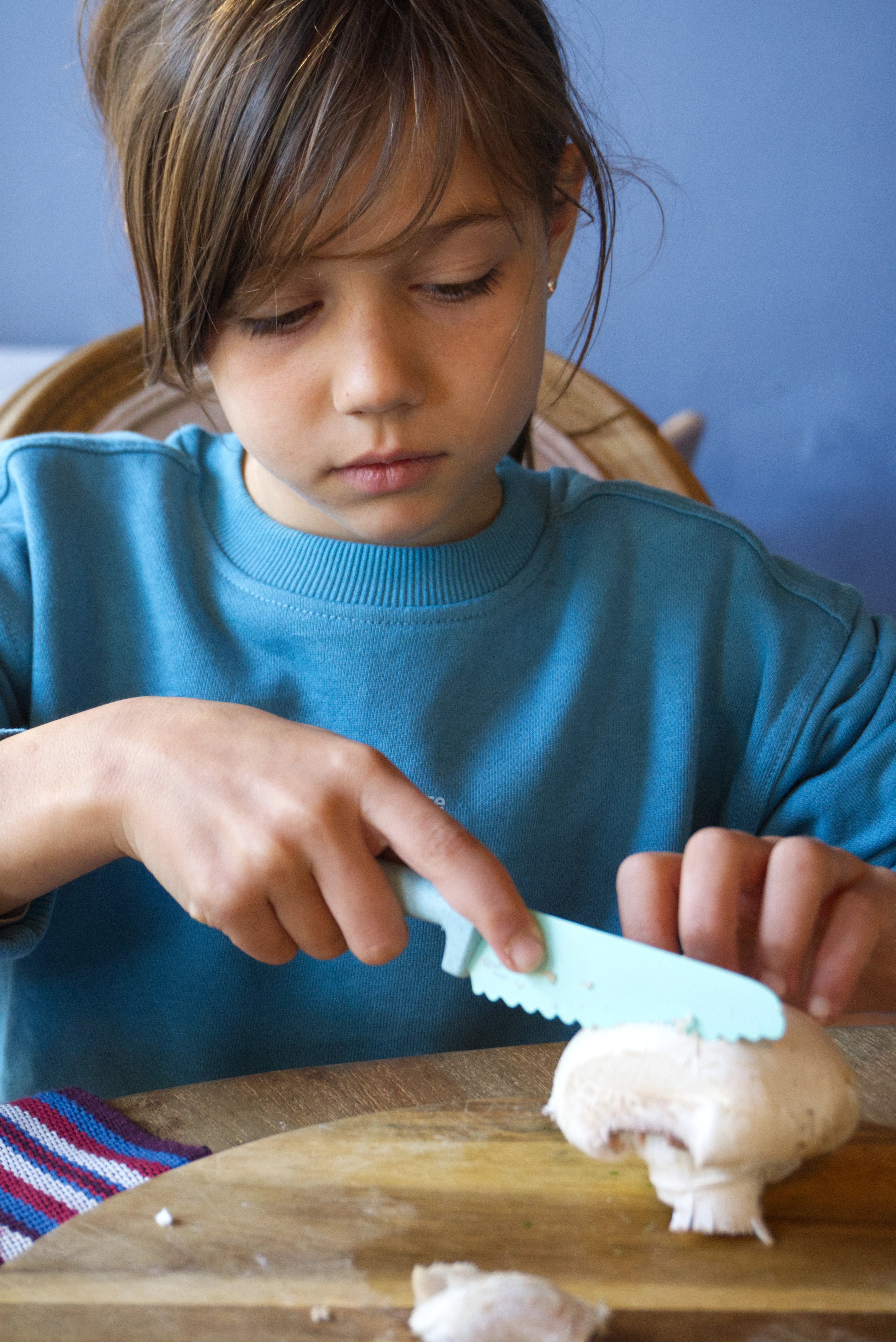 Child in a blue jumper cutting a mushroom on a wooden cutting board. Zara kids Sweater , Klyv classic knife for 3 years old Scandinavian Design, Skagfa