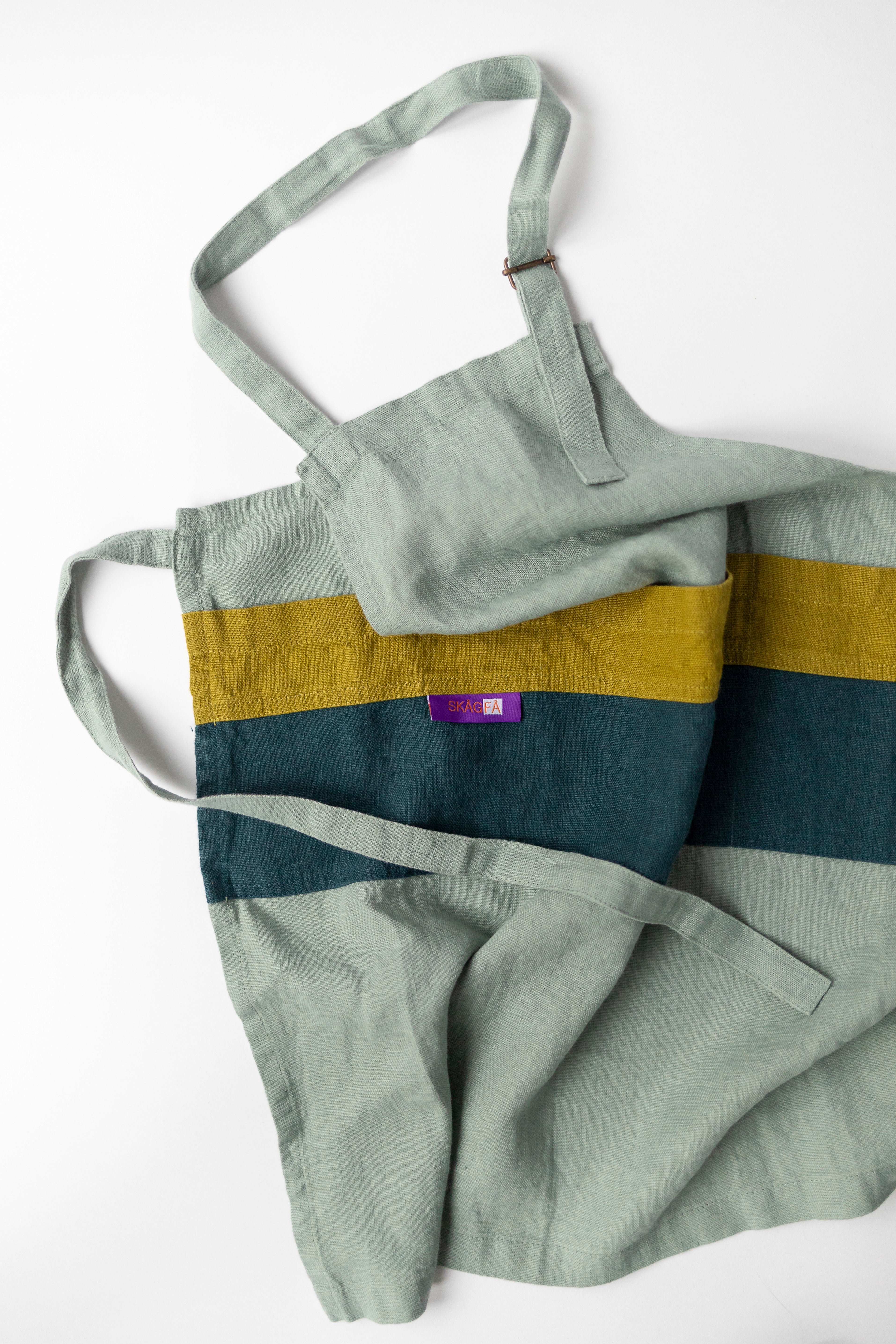 Child in a kitchen wearing 'The Förkläde' linen chef apron by SKÅGFÄ, showcasing adjustable neck strap and deep pockets, designed in Spain and handmade in Europe, perfect for culinary adventures. The apron features chic simplicity with 100% OEKO-TEX certified stone-washed linen, promoting sustainable fashion and functionality for young chefs blue navy pink peach salmon sage green forest