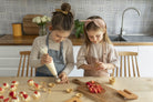 Child in a kitchen wearing 'The Förkläde' linen chef apron by SKÅGFÄ, showcasing adjustable neck strap and deep pockets, designed in Spain and handmade in Europe, perfect for culinary adventures. The apron features chic simplicity with 100% OEKO-TEX certified stone-washed linen, promoting sustainable fashion and functionality for young chefs blue navy pink peach salmon sage green forest. strawberries