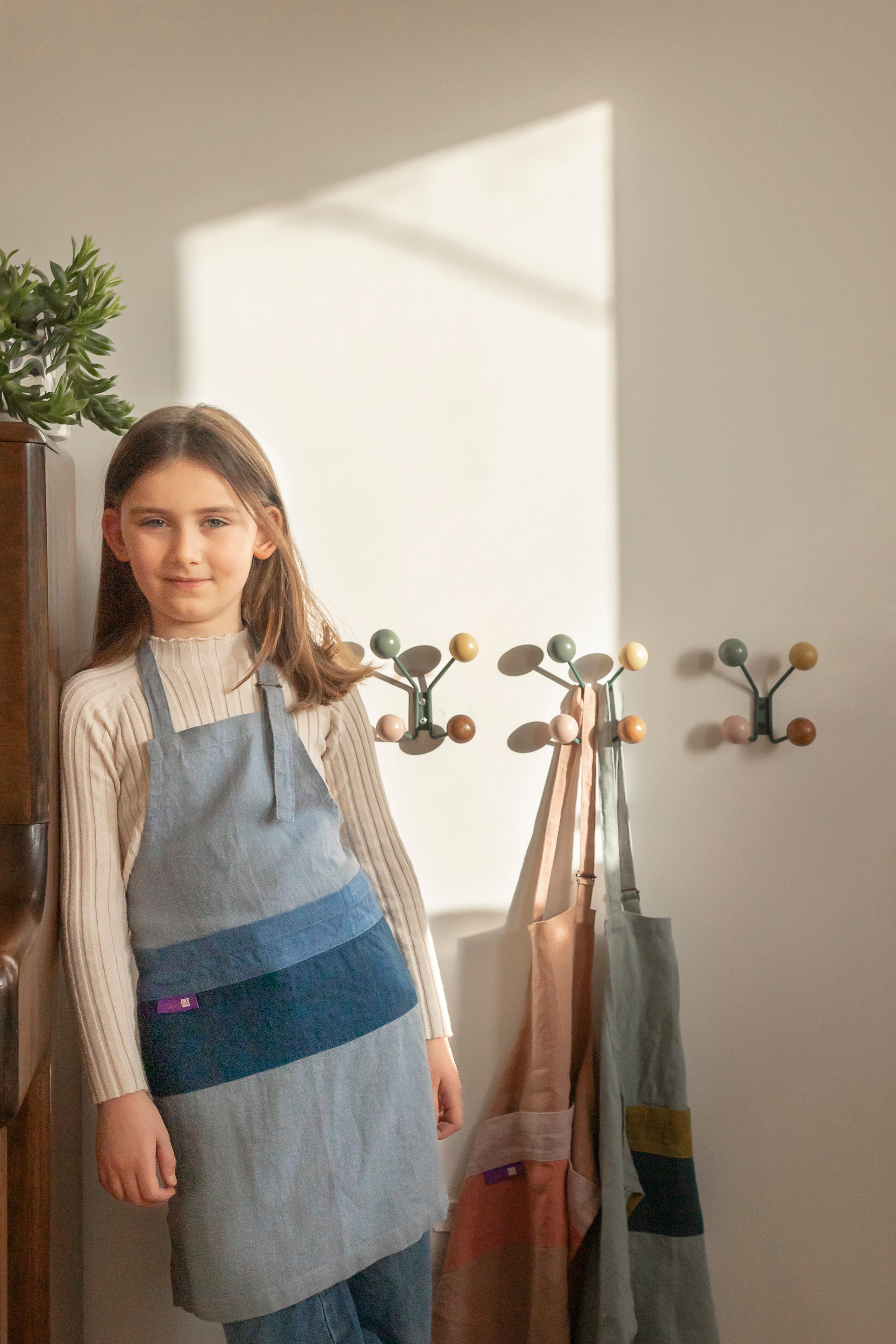 Child in a kitchen wearing 'The Förkläde' linen chef apron by SKÅGFÄ, showcasing adjustable neck strap and deep pockets, designed in Spain and handmade in Europe, perfect for culinary adventures. The apron features chic simplicity with 100% OEKO-TEX certified stone-washed linen, promoting sustainable fashion and functionality for young chefs blue navy pink peach salmon sage green forest